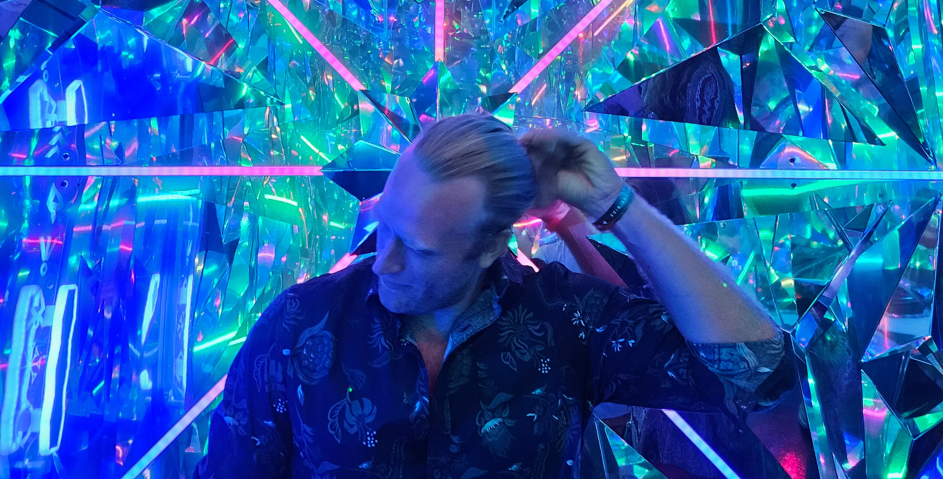 Scott Veltkamp inside a mirrored, crystalline art installation lit in blue, green and pink neon, one hand raised to his hair.